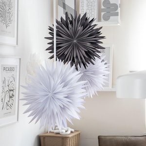 Puede incluir: Tres adornos de estrellas de papel colgando del techo. Las estrellas son blancas y grises y están hechas de papel doblado. Las estrellas están colgando en una sala de estar con una mesa de madera y una silla de cuero marrón.