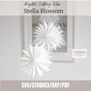 Könnte beinhalten: Zwei weiße Papiersterne mit spitzen Blütenblättern, die an dünnen Schnüren hängen. Der Text lautet Digital Cutting Files Stella Blossom, VIDEO TUTORIAL und SVG/STUDIO3/DXF/PDF. Diese geometrischen Papiersterne haben ein radiales Faltmuster.