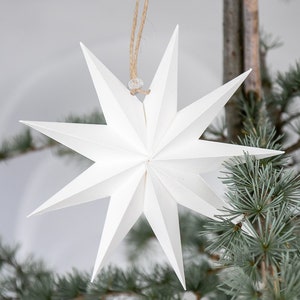 May include: A white, multi-pointed star ornament constructed from folded paper with sharp, geometric edges. The star features a central point and radiates outward into eight distinct, symmetrical points. It hangs from a thin, natural jute twine loop.