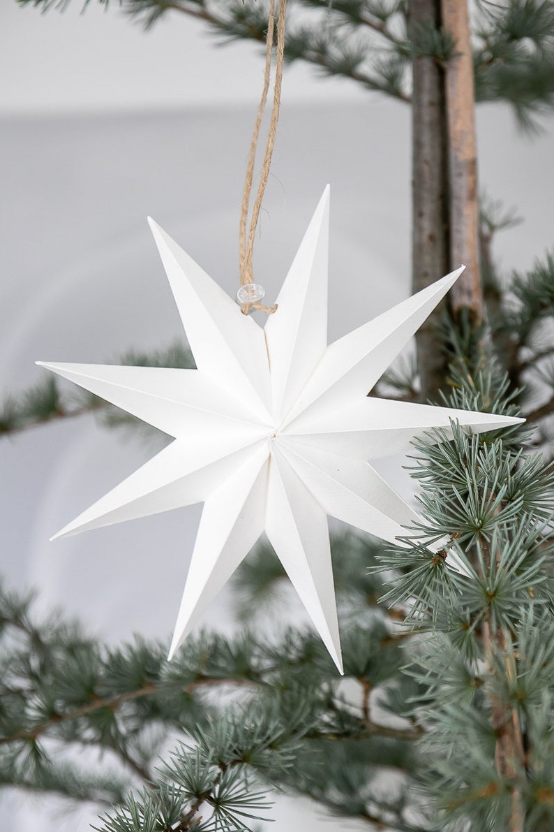 May include: A white, multi-pointed star ornament constructed from folded paper with sharp, geometric edges. The star features a central point and radiates outward into eight distinct, symmetrical points. It hangs from a thin, natural jute twine loop.