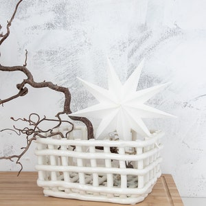 May include: A white ceramic basket with a woven, grid-like structure. Inside the basket sits a white, multi-pointed star ornament and a dark, twisted, leafless branch that extends outward from the center of the basket's open top.