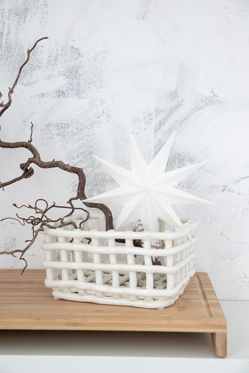 May include: A white ceramic basket with a woven, grid-like structure. Inside the basket sits a white, multi-pointed star ornament and a dark, twisted, leafless branch that extends outward from the center of the basket's open top.