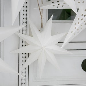 May include: A collection of white paper star ornaments hanging from brown twine strings. The stars feature sharp, multi-pointed designs, with some variations displaying a pattern of small, circular cutouts across their surfaces.