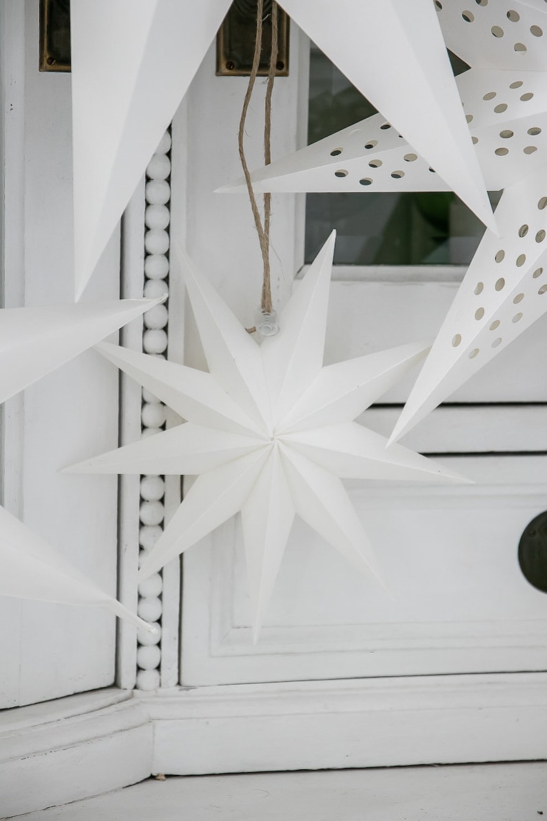 May include: A collection of white paper star ornaments hanging from brown twine strings. The stars feature sharp, multi-pointed designs, with some variations displaying a pattern of small, circular cutouts across their surfaces.