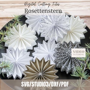Op de afbeelding: Een verzameling gevouwen papieren sterornamenten in witte, grijze en zwarte patronen, gerangschikt op een zwart oppervlak met dennentakjes. De tekst luidt: Digital Cutting Files Rosettenstern, VIDEO TUTORIAL en SVG/STUDIO3/DXF/PDF.