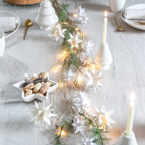 Op de afbeelding: Een witte tafelsetting met een wit tafelkleed, een witte keramische kerstboom kaarsenhouder, een witte keramische ster kaarsenhouder en een witte keramische kom gevuld met koekjes. De tafel is versierd met een krans van groenblijvende takken en witte papieren sterren, en een string witte lampjes.