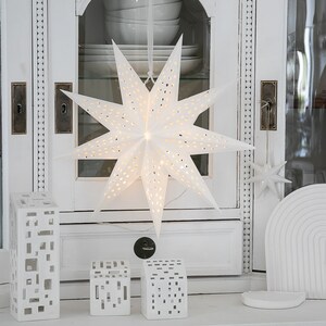 May include: A white paper star lantern with perforated holes hanging from a ribbon, accompanied by three white ceramic house-shaped candle holders with rectangular cutouts and a small white star ornament hanging to the right side.
