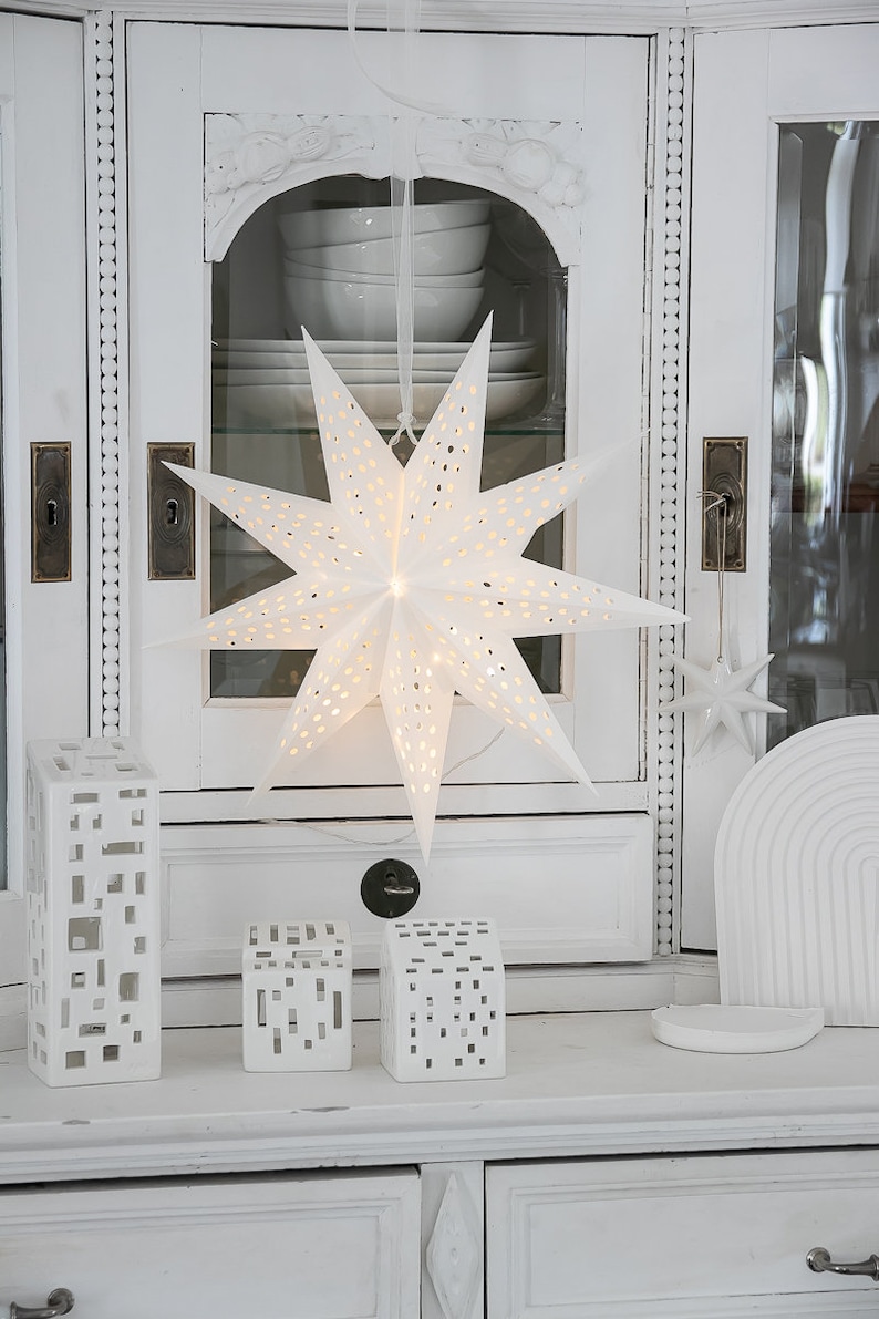 May include: A white paper star lantern with perforated holes hanging from a ribbon, accompanied by three white ceramic house-shaped candle holders with rectangular cutouts and a small white star ornament hanging to the right side.