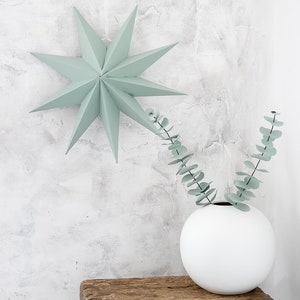 May include: A sage green paper star ornament with multiple points hangs above a white spherical vase containing two stems of eucalyptus leaves. The vase rests on a rustic wooden stool with a weathered texture and visible wood grain patterns.