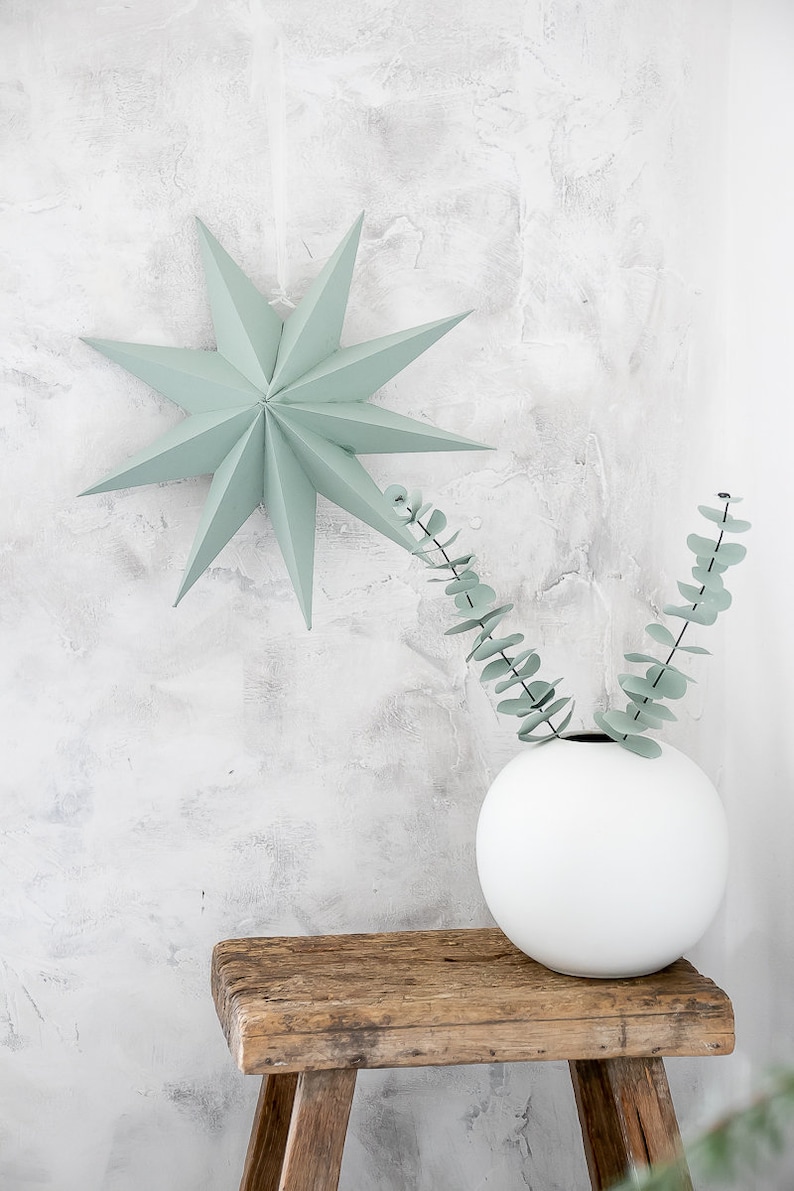 May include: A sage green paper star ornament with multiple points hangs above a white spherical vase containing two stems of eucalyptus leaves. The vase rests on a rustic wooden stool with a weathered texture and visible wood grain patterns.