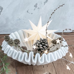 May include: A white, round, fluted bowl containing a large, multi-pointed white star, several brown pinecones, and a string of small, illuminated star-shaped lights intertwined with thin, bare branches and green pine needles.