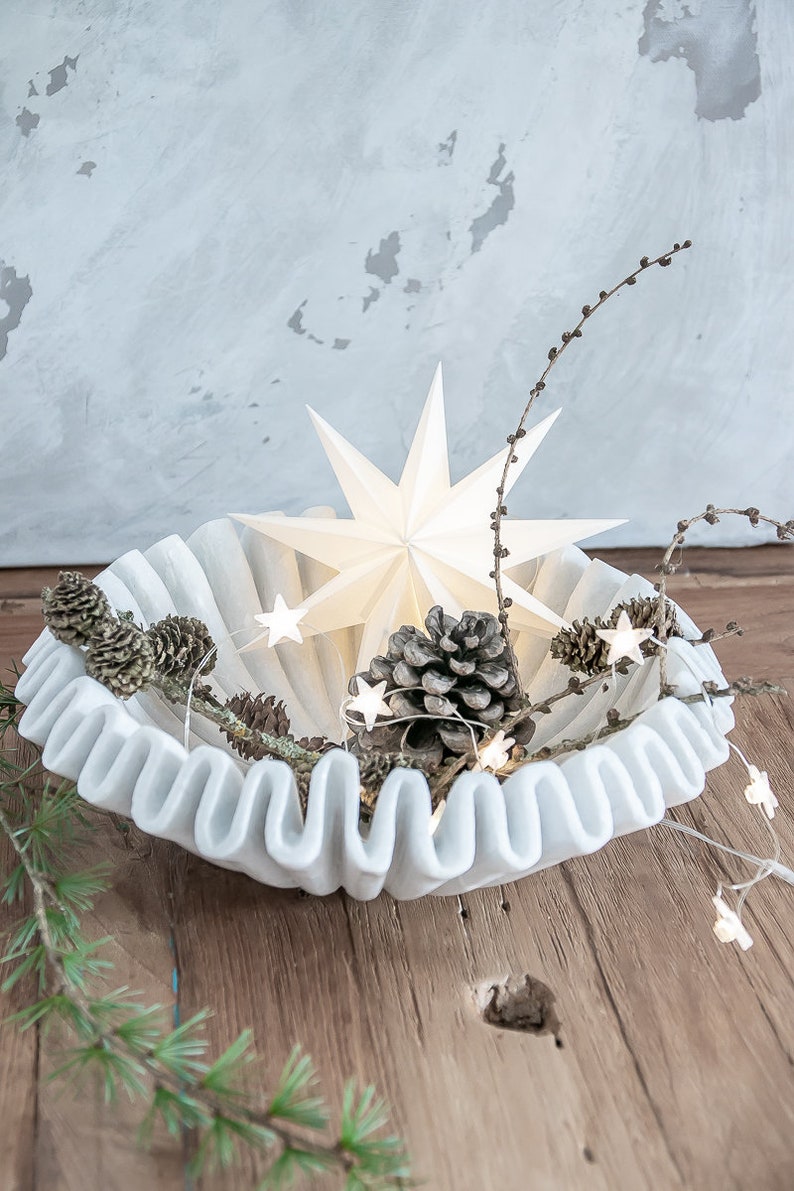 May include: A white, round, fluted bowl containing a large, multi-pointed white star, several brown pinecones, and a string of small, illuminated star-shaped lights intertwined with thin, bare branches and green pine needles.