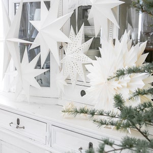 May include: A collection of white paper star ornaments in various shapes and sizes, including multi-pointed stars and one with a perforated pattern. These decorative hanging stars are displayed in front of a white cabinet with glass doors.