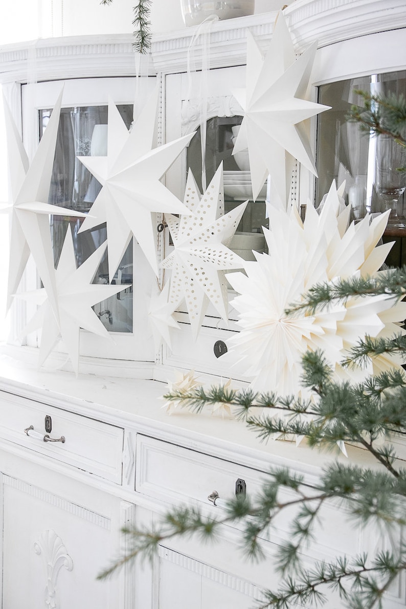 May include: A collection of white paper star ornaments in various shapes and sizes, including multi-pointed stars and one with a perforated pattern. These decorative hanging stars are displayed in front of a white cabinet with glass doors.