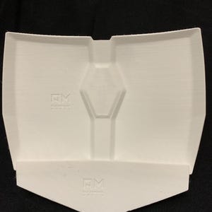 Op de afbeelding: Een wit, 3D-geprint kunststof onderdeel met een centrale zeshoekige inkeping en een apart, kleiner, hoekig wit stuk aan de onderkant. Beide onderdelen tonen het in reli&euml;f gemaakte logo NM met tekst eronder op hun oppervlakken.