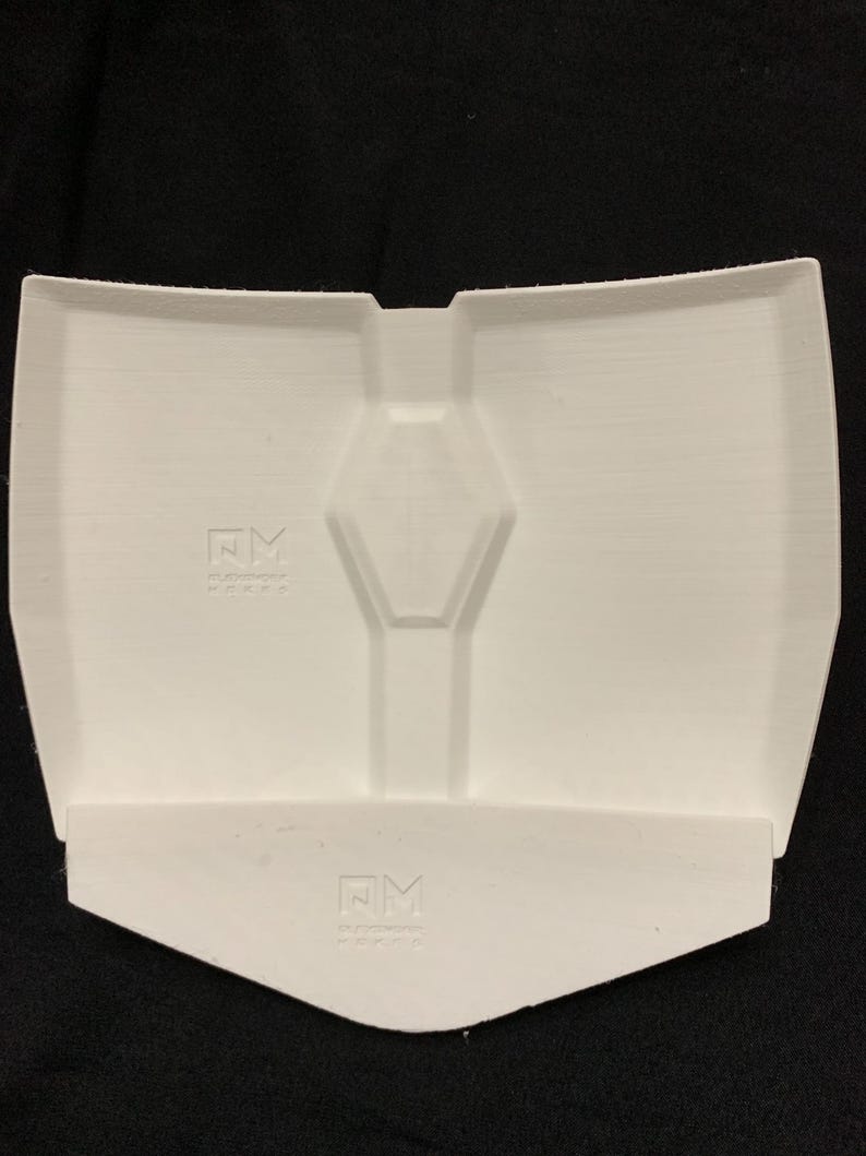 Op de afbeelding: Een wit, 3D-geprint kunststof onderdeel met een centrale zeshoekige inkeping en een apart, kleiner, hoekig wit stuk aan de onderkant. Beide onderdelen tonen het in reli&euml;f gemaakte logo NM met tekst eronder op hun oppervlakken.