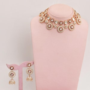 Puede incluir: Un collar dorado y un juego de pendientes a juego exhibidos en un maniquí rosa. El collar y los pendientes presentan piedras preciosas rojas y transparentes. Dos flores decorativas se encuentran en la base.