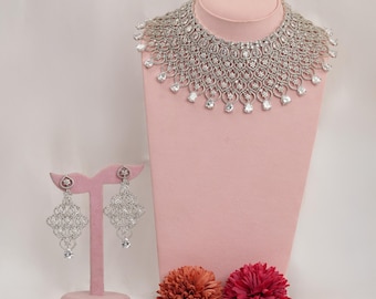 May include: A dazzling silver-toned necklace and matching earrings set. The necklace features an intricate design with numerous clear, sparkling stones. The earrings are a similar design, with a dangling stone at the bottom. The jewelry is displayed on a pink mannequin.