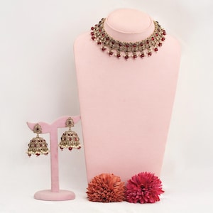Può includere: Un set di gioielli color oro composto da una collana girocollo e orecchini abbinati. La collana e gli orecchini sono adornati con pietre preziose rosse e piccole perle bianche. I gioielli sono esposti su un manichino e un supporto rosa, con due fiori decorativi alla base.