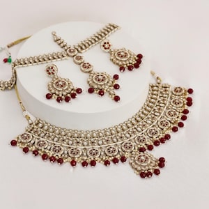 Op de afbeelding: Een goudkleurige sieradenset met een ketting, oorbellen en een hoofdband. De set is versierd met kleine witte kralen en rode accenten. De ketting en oorbellen hebben een bloemmotief met bungelende rode kralen.
