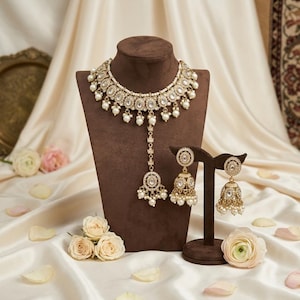 Puede incluir: Un conjunto de collar y pendientes en tono dorado con perlas artificiales blancas y cristales transparentes. El collar tiene un colgante central y los dos pendientes a juego son de estilo jhumka con forma de campana y detalles de perlas.
