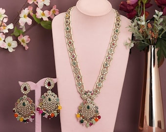May include: A jewelry set featuring a long necklace and matching earrings. The necklace and earrings are adorned with colorful gemstones and pearls. The set is displayed on a pink mannequin, with a vase of flowers in the background.