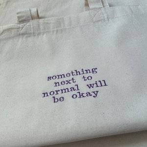 Pode incluir: Várias sacolas de lona branca com alças curtas estão empilhadas. A frase "something next to normal will be okay" está impressa em roxo na frente de uma sacola. As sacolas são feitas de um tecido resistente.