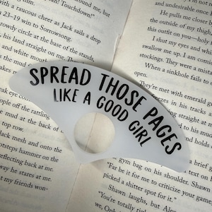 Puede incluir: Un sujetalibros de plástico blanco translúcido con forma de abanico y un orificio circular en el centro. El texto SPREAD THOSE PAGES LIKE A GOOD GIRL está impreso en letras mayúsculas negras y negritas sobre la superficie del accesorio.