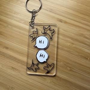 May include: A clear acrylic keychain with a silver keyring and chain. The keychain features two white speech bubbles with the word "HI" in black, and black leaf-like designs. The background is a wooden surface.
