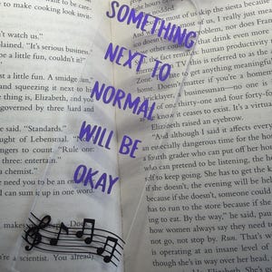 Può includere: Segnalibro trasparente con testo viola: "SOMETHING NEXT TO NORMAL WILL BE OKAY." Presenta un pentagramma con note musicali. Include un nastro rosa. Posizionato su un libro aperto.