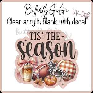 May include: A clear acrylic blank with a UV-DTF decal featuring the text 'TIS' THE SEASON' in black script. The design includes an iced coffee with a pink bow, a football, a plaid helmet, a pumpkin, and autumn leaves on a light pink background.
