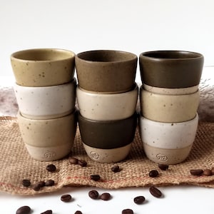 Könnte beinhalten: Neun kleine, handgefertigte Espressotassen aus Keramik in verschiedenen Beige-, Braun- und Olivgrüntönen. Die Tassen sind in drei Stapeln angeordnet. Die Tassen haben eine gesprenkelte Textur und einen abgerundeten Boden. Kaffeebohnen liegen verstreut.