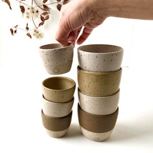 Pode incluir: Uma coleção de tigelas de cerâmica feitas à mão em várias cores terrosas, incluindo bege, castanho claro e castanho. As tigelas têm uma textura salpicada e um design minimalista, perfeitas para servir comida ou como peças decorativas. Uma mão segura uma das tigelas.