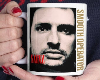 May include: White ceramic mug with a black and white photo of a man's face. The text "SAINZ" is in red letters at the bottom of the photo. The text "SMOOTH OPERATOR" is in gold letters on the right side of the mug.
