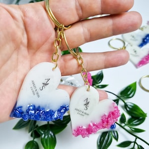 May include: Two white heart-shaped keychains with gold chains. The keychains have blue and pink glitter embedded in the bottom half of the heart. The top half of the heart has the initials "A & S" and the text "Ashley & Sean 2.2.2023".