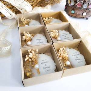 May include: Small, white, round, favor boxes with a clear top. Each box contains a small bouquet of dried white flowers, a pearl, and a white card with the text "Thank You Vanessa & Daniel 18.08.2023".