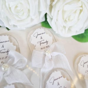 May include: Clear acrylic wedding favors with white text that reads "Jeanine & Frandy" and the date "06.12.2025". Each favor is adorned with a white ribbon and a pearl. White roses are in the background.