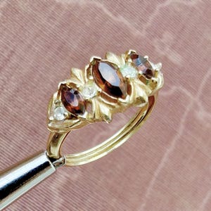 May include: A gold ring featuring three marquise-cut dark red gemstones arranged horizontally, interspersed with small, round, clear accent stones. The metal setting has a textured, leaf-like design surrounding the stones on a polished gold band.