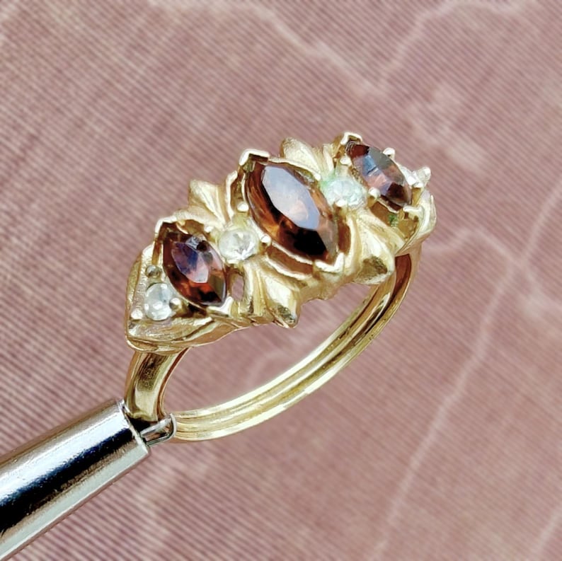 May include: A gold ring featuring three marquise-cut dark red gemstones arranged horizontally, interspersed with small, round, clear accent stones. The metal setting has a textured, leaf-like design surrounding the stones on a polished gold band.
