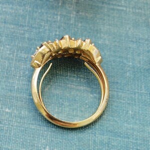 May include: A gold ring featuring a textured, multi-tiered top section with a curved, polished band. The design displays a series of small, raised, metallic segments arranged in a cluster, creating a layered appearance on the upper portion of the ring.