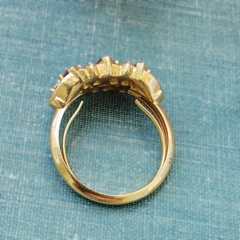 May include: A gold ring featuring a textured, multi-tiered top section with a curved, polished band. The design displays a series of small, raised, metallic segments arranged in a cluster, creating a layered appearance on the upper portion of the ring.