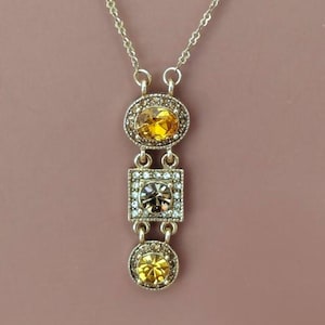 May include: A gold-toned pendant necklace with three jewel-encrusted charms. The top charm is an oval shape, the middle is square, and the bottom is round. Each charm features a large, central stone surrounded by smaller, clear stones.