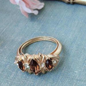May include: A gold-toned ring featuring a bypass band design. The centerpiece is adorned with three marquise-cut brown gemstones and two small clear round stones, all set within a textured, decorative metal mounting with a polished finish.