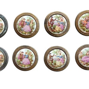 May include: A set of eight circular buttons featuring metallic gold-toned rims with a ribbed texture. Each button contains a central ceramic-like inlay depicting colorful illustrations of figures in historical attire within outdoor garden settings.