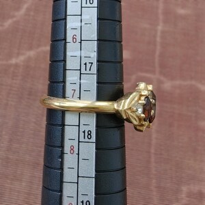 May include: A gold ring featuring a dark brown oval gemstone flanked by two small clear stones, positioned on a black ring mandrel with a visible scale showing a size between 17 and 18 cm on the metric measurement side of the tool.
