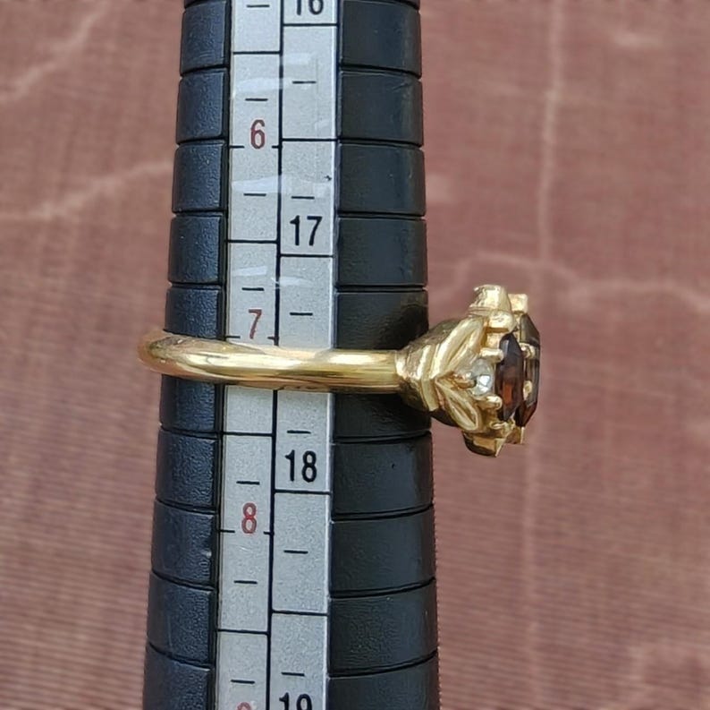 May include: A gold ring featuring a dark brown oval gemstone flanked by two small clear stones, positioned on a black ring mandrel with a visible scale showing a size between 17 and 18 cm on the metric measurement side of the tool.