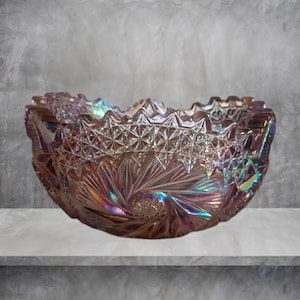 May include: Iridescent glass bowl with a starburst and geometric pattern. The bowl has a ruffled edge and a shimmering, multi-colored finish, reflecting hues of purple, blue, and gold. The bowl is likely vintage or antique.
