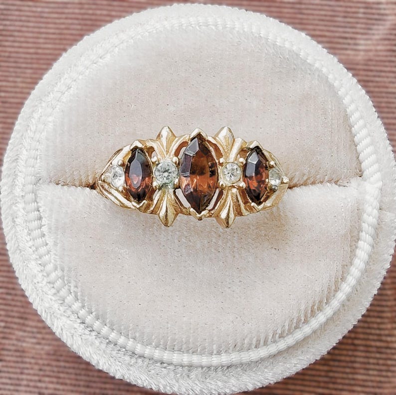 May include: A gold ring featuring three marquise-cut brown gemstones arranged horizontally, accented by four small round clear stones set between the larger gems. The metal band has a decorative, pointed prong setting holding each stone securely in place.