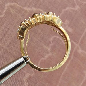 May include: A gold ring featuring a curved row of multiple small, clear, faceted stones set along the top band. The metal band is polished and circular, showing a side profile view of the setting where the stones are secured in individual prongs.