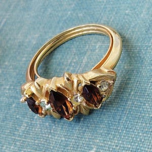 May include: A gold-toned ring featuring three marquise-cut dark brown stones arranged horizontally across the front. The band is smooth and polished, accented by small, clear, round stones nestled between the larger brown marquise-cut gems.